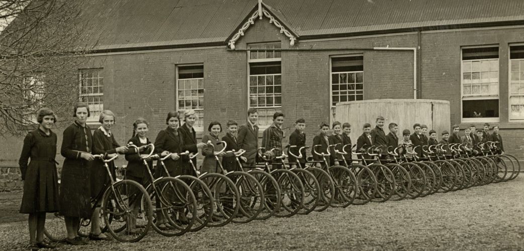 An old black and white photo of a line of school kids and their bikes.