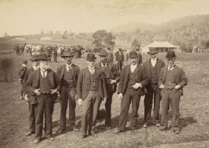 An old black and white photo of 8 men posing for a photo. a crowd of people and a horse in the background.