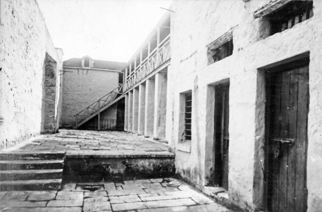 A black and white photo of an enclosed outdoor area made of brick. stairs leading to a second level are at the end.