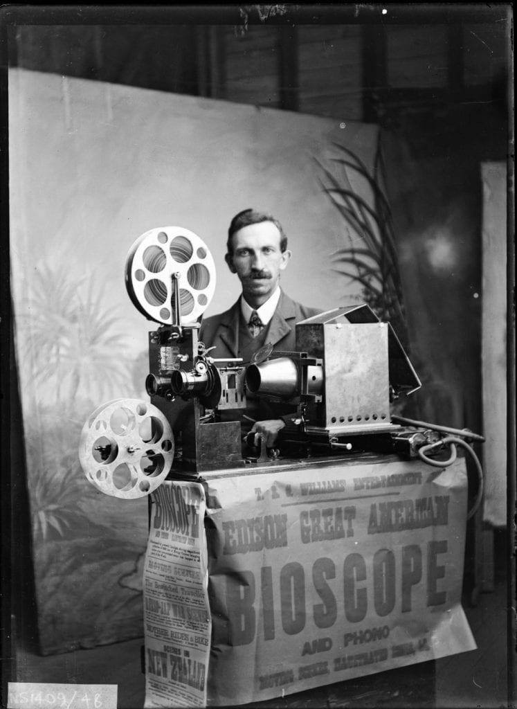 A black and white photo of a man standing behind an old film projector.