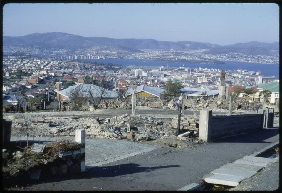 Rubble where buildings once stood. Seen from the road atop a hill. untouched houses in the distance.