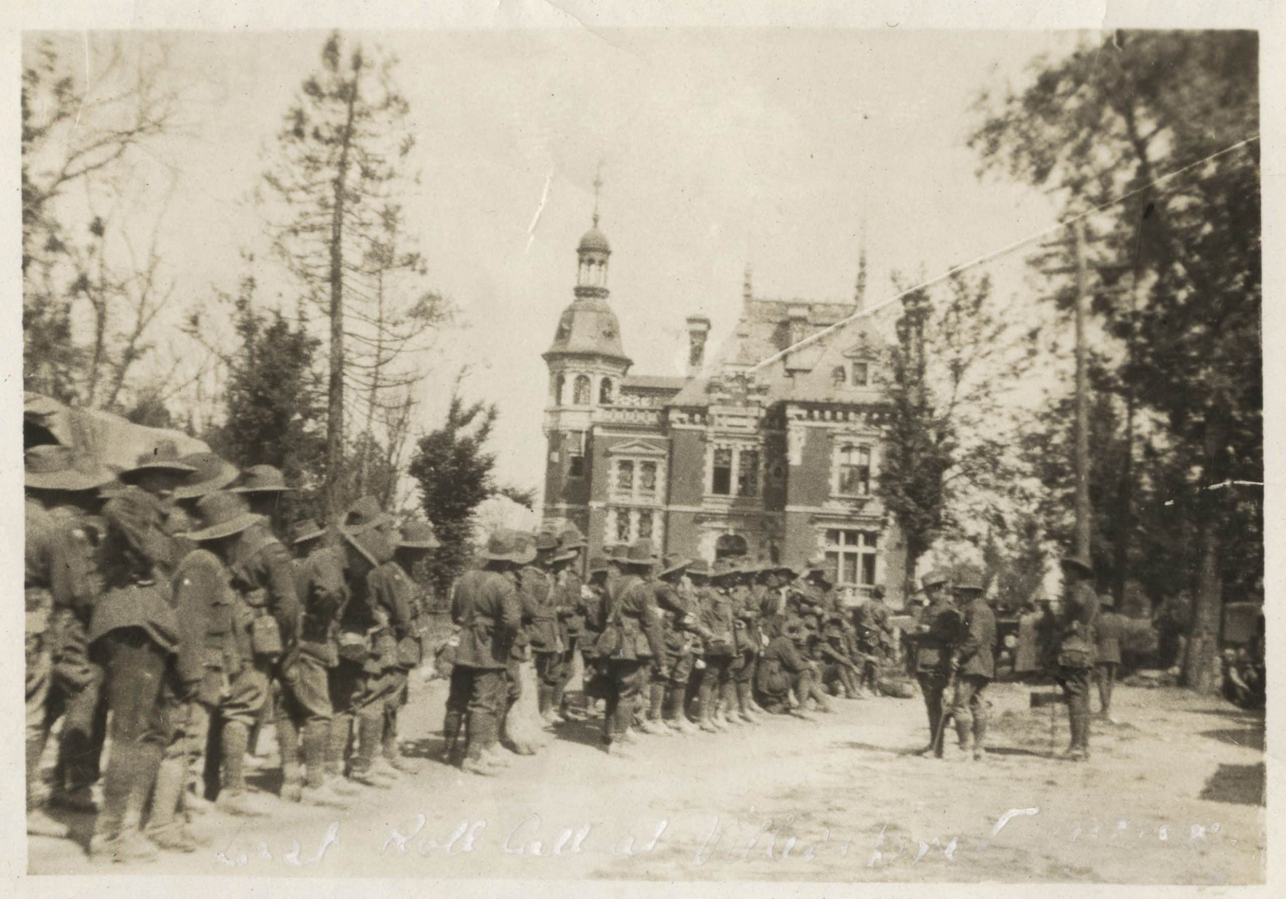 Black and white photo of world war one soldiers along the edge of a road awaiting instructions from the officers standing in the middle of the road. In the background is a chateau.