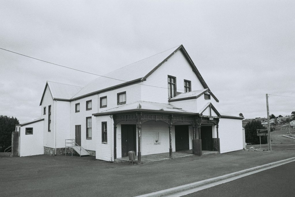 A black and white photo of a white, two-story weather board building on the side of a main road.