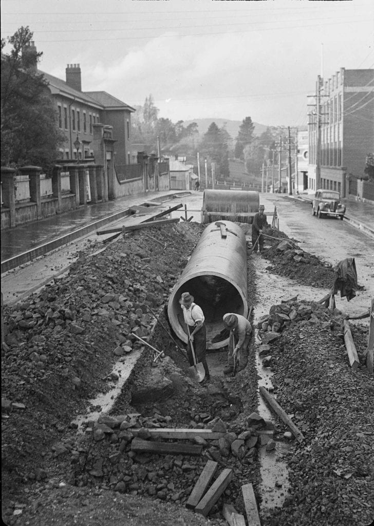 2 workmen working on a road. they are digging, a Large pipe sits in a trench in the road.