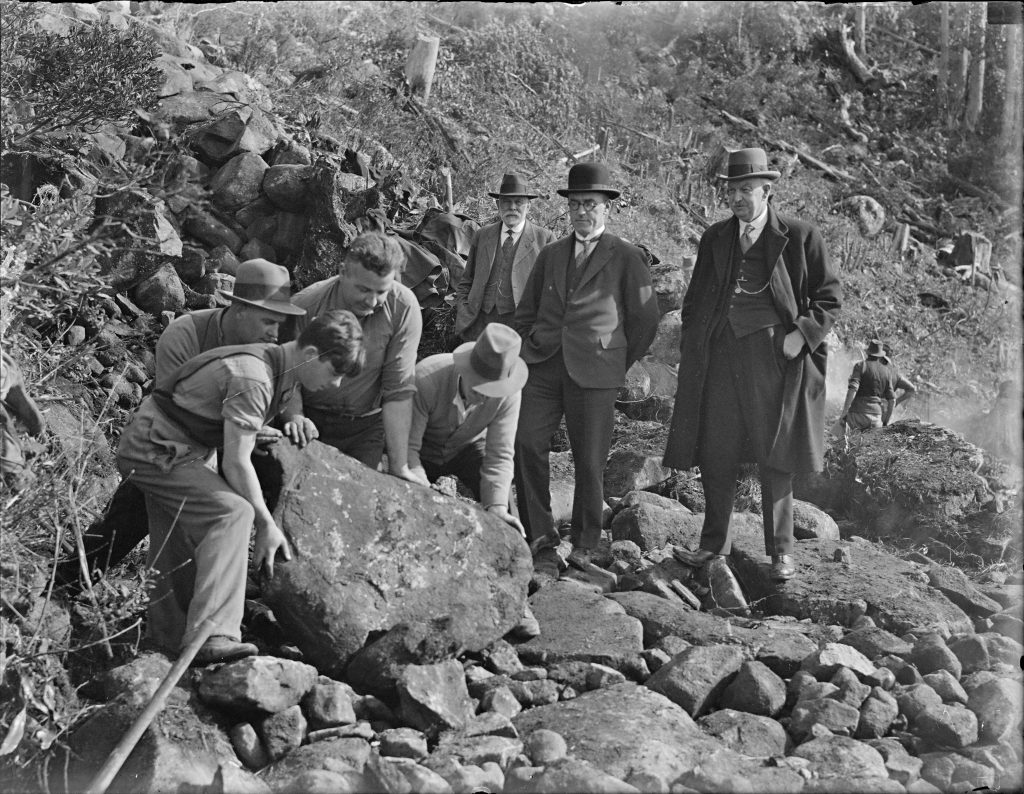 An old black and white photo of 3 men in suits standing watching 4 other men move a large rock.