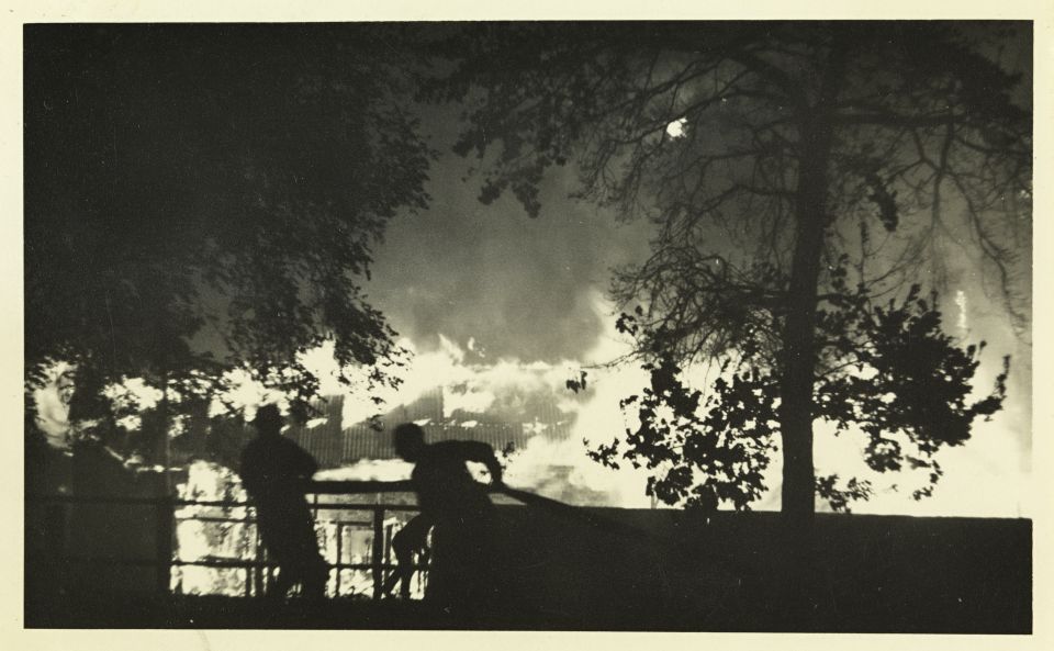A black and white photo of people standing in front of a fence watching buildings on fire.