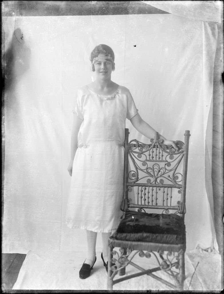 An old black and white photo of a woman standing next to a chair.