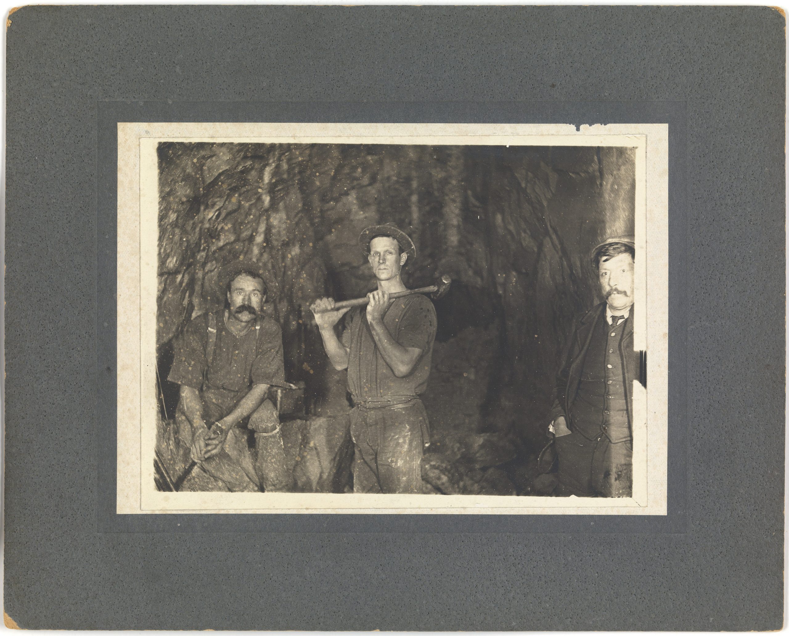 An old black and white photo of 3 men inside a mine. One is holding a sledgehammer.