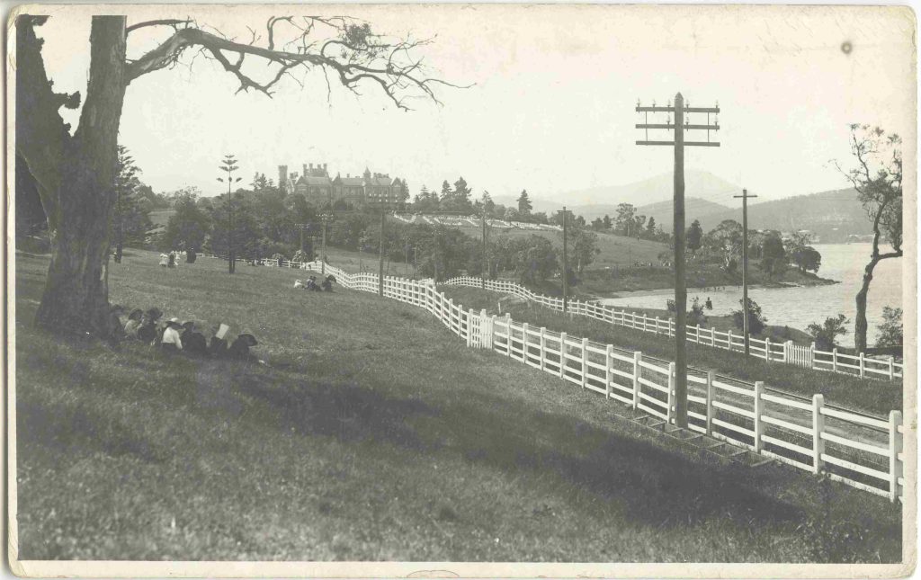 A black and white photo of a road along a grassy hill beside a river. People picnic on the grass. There is a large home on a hill in the background. The word "victory" has been laid out on the grass in front of the home.