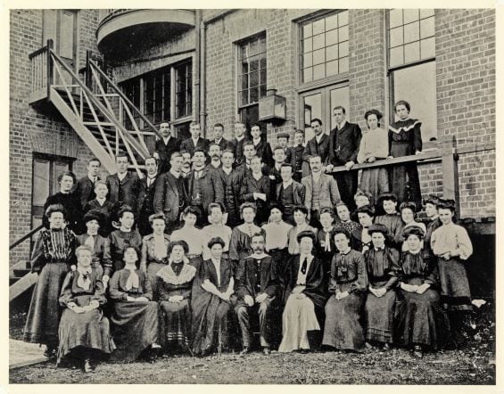 A black and white group photo of about 40 people standing in front of a brick building.
