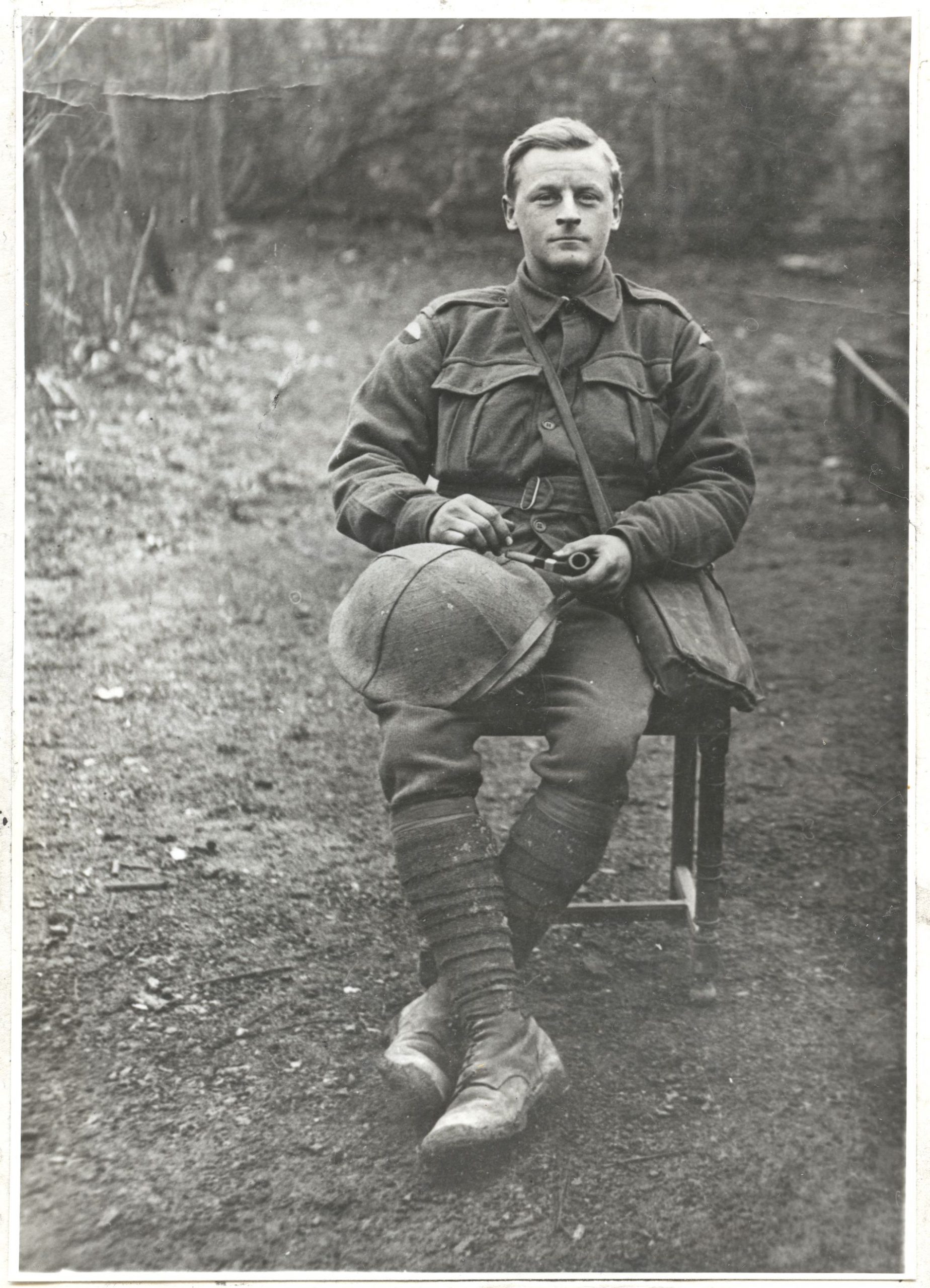 A black and white photo of a man dressed in a world war one soldiers uniform. He is sitting on a chair outside holding onto his tobacco pipe and helmet.
