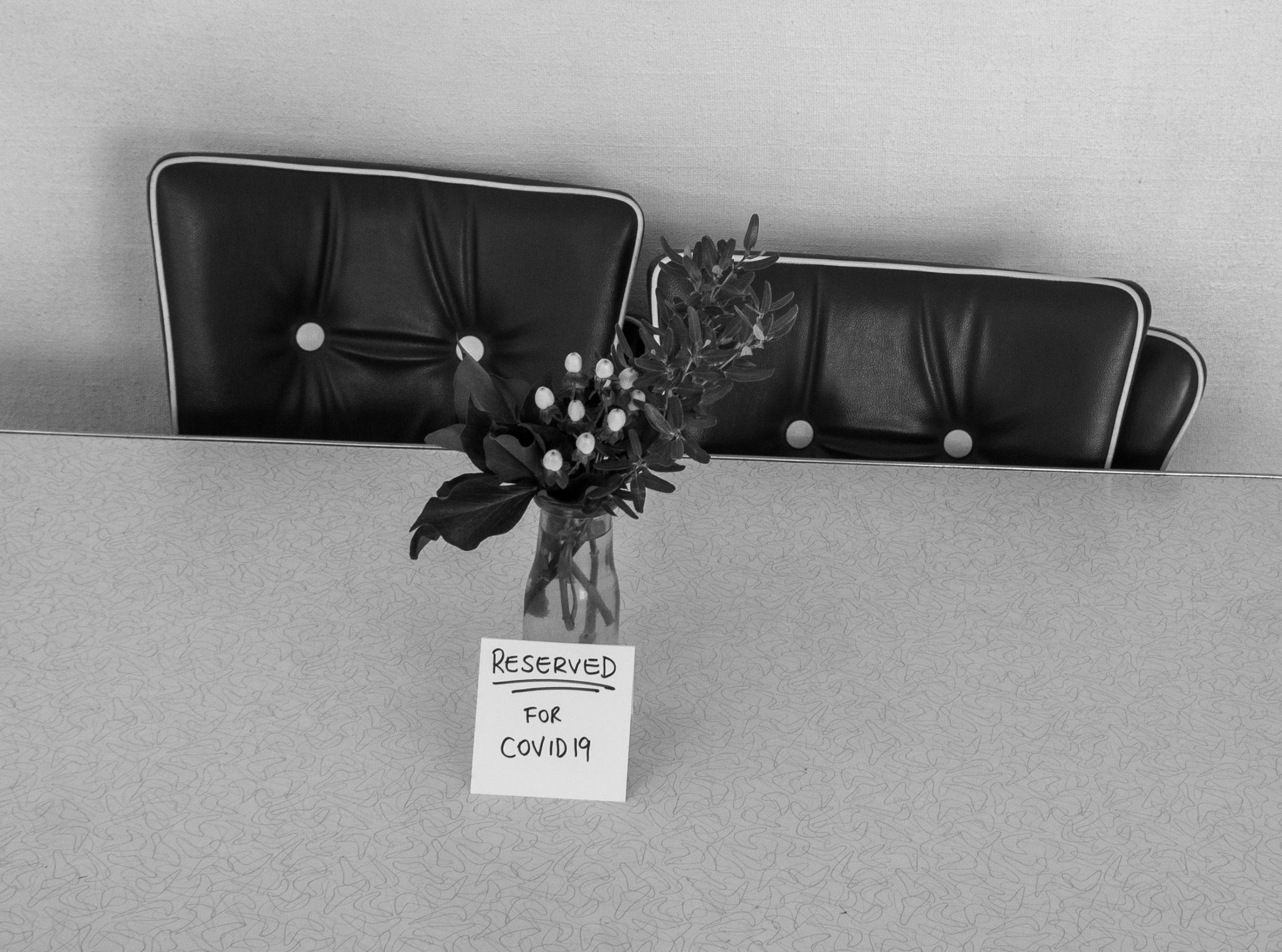 A black and white photo. Flowers in a vase on a table. a sign on the table reads: "Reserved for Covid19"
