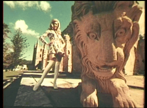 a still taken from Film – The Other Face Of The Island. A woman standing next to a lion statue