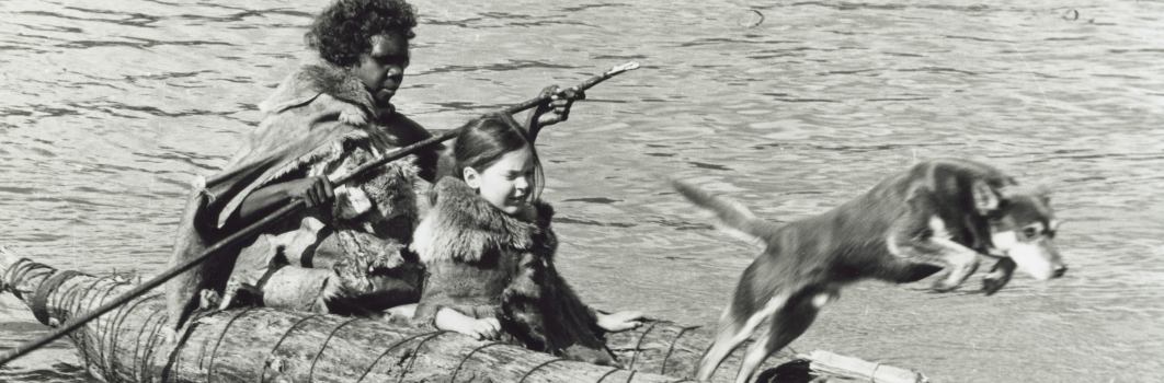 A still image from a film. A woman, a girl and a dog on a raft. The dog is jumping into the water.