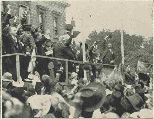 A black and white photo of a crowd of people cheering in front of a building. Officials stand on a platform in front of the crown.