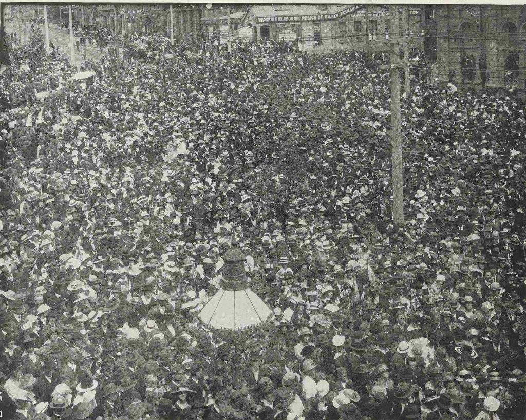 A black and white photo of a crowd of people in the street, taken from above looking back down the street. Power poles and lines can be seen on the edges of the street. In the background are the buildings on the other side of the street.