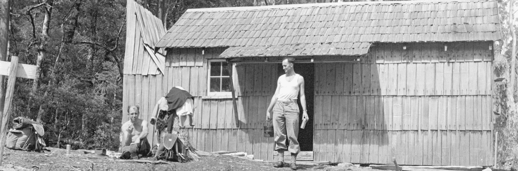 A man sitting and a man standing out the front of a wooden building. Dense trees in the background.