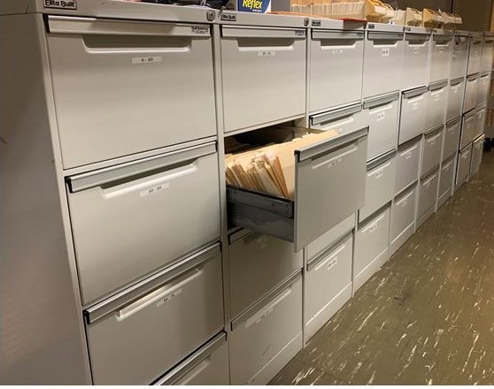 A row of fiing cabinets. one drawer is open and contains manilla folders.