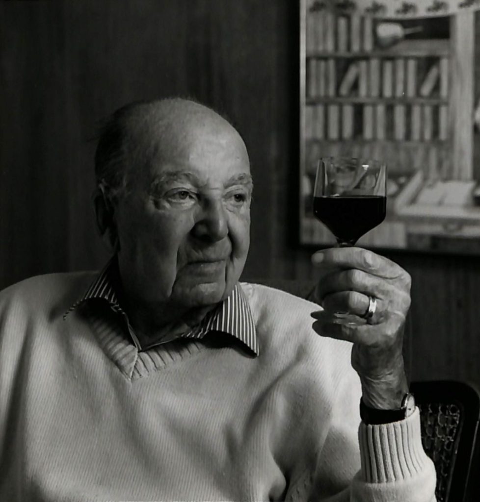 Black and white portrait photo of an elderly man neatly dressed in a business shirt and light-coloured. He is studying a glass of wine held in his right hand. He is sitting down in a show room.
