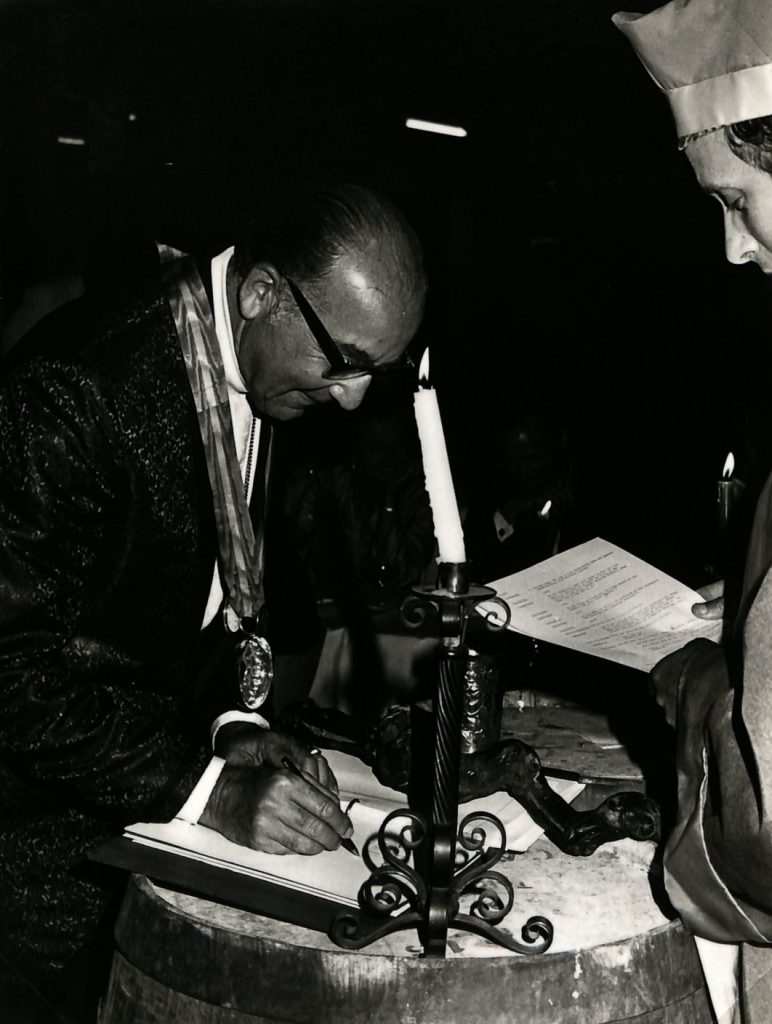 A black and white photo of a man at a formal dinner.  He is standing up beside a wine barrel which he uses to lean on while signing papers.