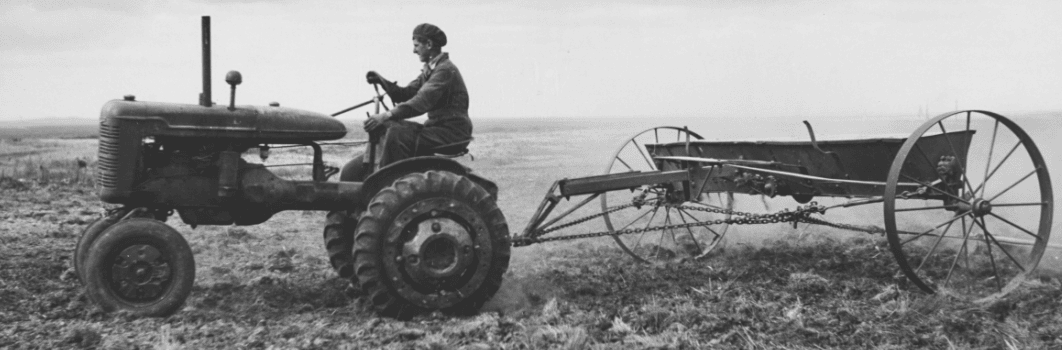A black and white photo of a man on a tractor pulling a trailer in a field.