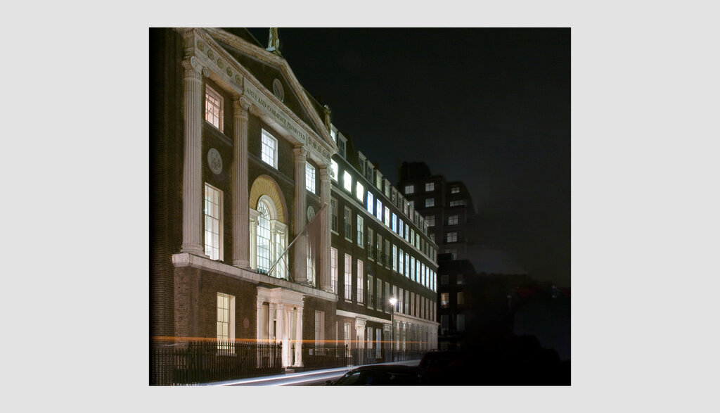 Nighttime view of the RSA House, an old brick building with lit windows, featuring a prominent circular emblem above the entrance. Street lights illuminate the scene, casting soft reflections on the road. Nearby, darkened modern buildings are visible.