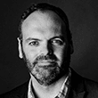 Black and white portrait of a man with a beard, wearing a suit jacket and a collared shirt. He has a calm expression and is facing slightly to the right. The background is dark and unobtrusive.