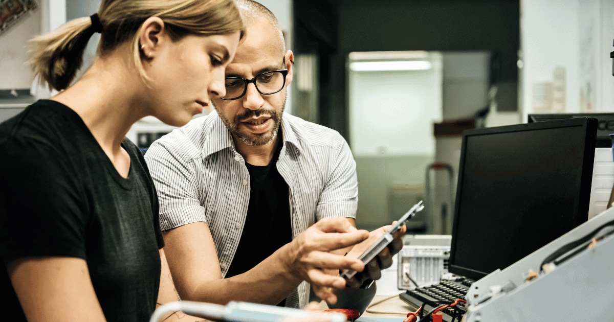Two people work in an office setting. One is showing a device to the other, who listens intently. A computer monitor is on the desk, surrounded by various electronic components and tools. The atmosphere is focused and collaborative.
