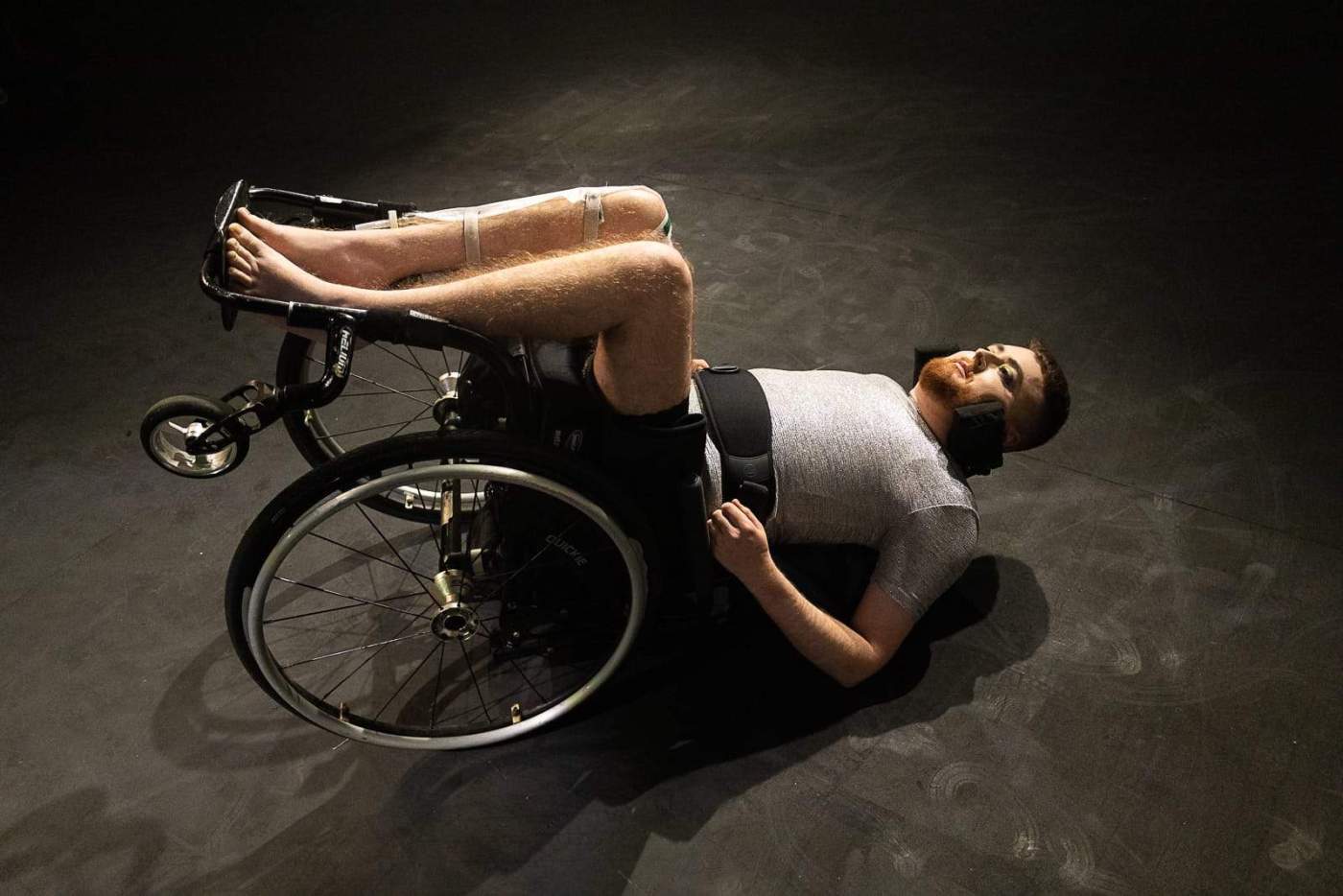 A person in a manual wheelchair, tipped over and lying on their back on the stage. It is a lightweight black manual wheelchair, containing Jamie, a white person with short dark red hair and a red beard, wearing a silver t-shirt, with a black band securing them to the chair round their waist. Their legs are bare, with catheter bag strips visible on one leg, and they are barefoot.