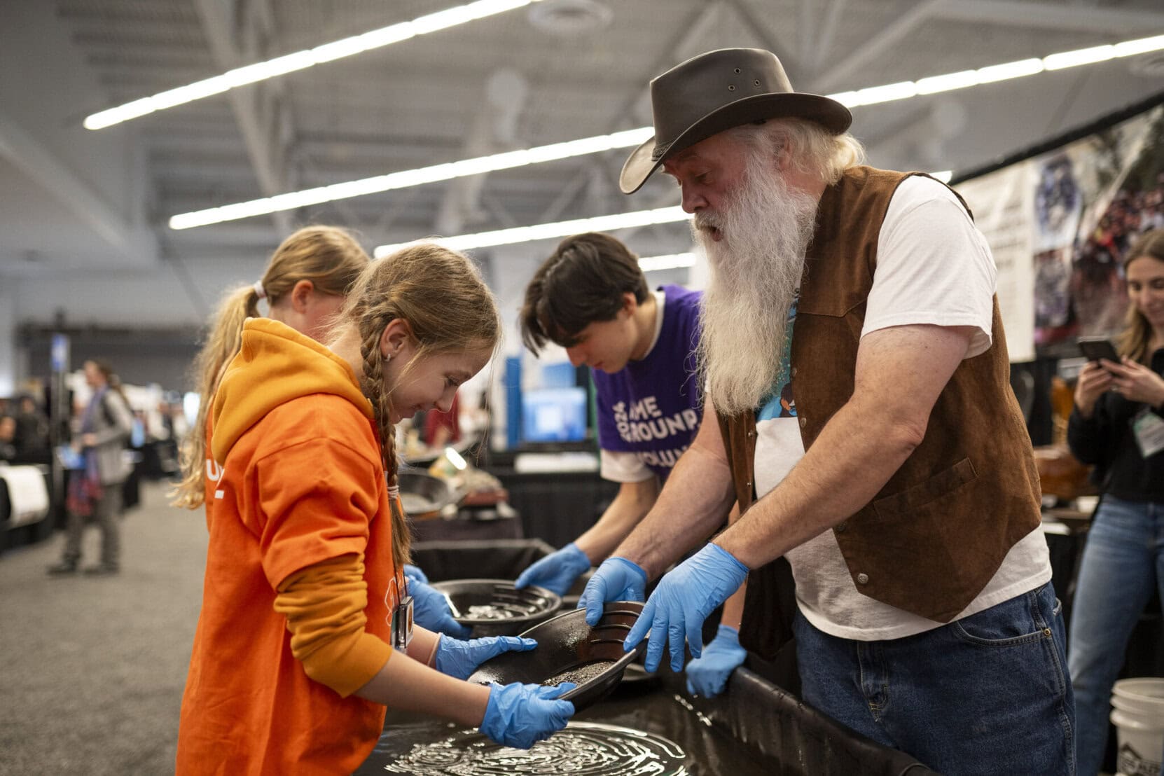 A man in a leather stetson and hide vest shows a young girl how to search a pan for gold at the AME conference