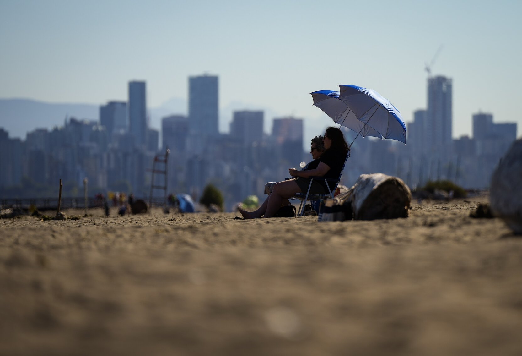 Two people find shade under an umbrella on an urban beach, with a hazy skyline behind