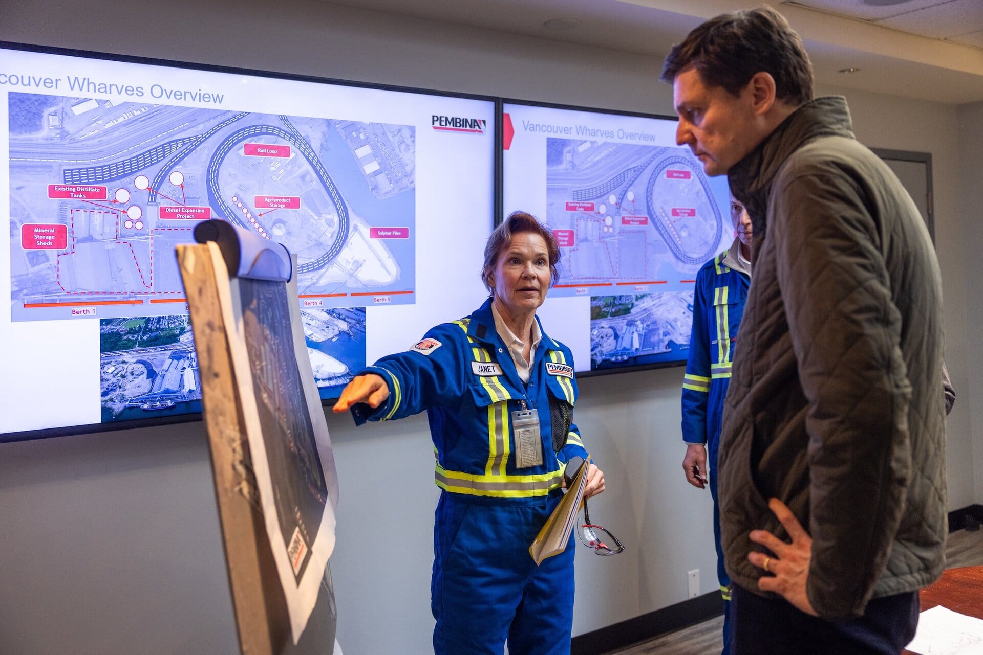 David Eby is speaking with a woman wearing blue coveralls who is pointing at a display board. Two large screens behind them show annotated aerial photos of the Vancouver Wharves export facility