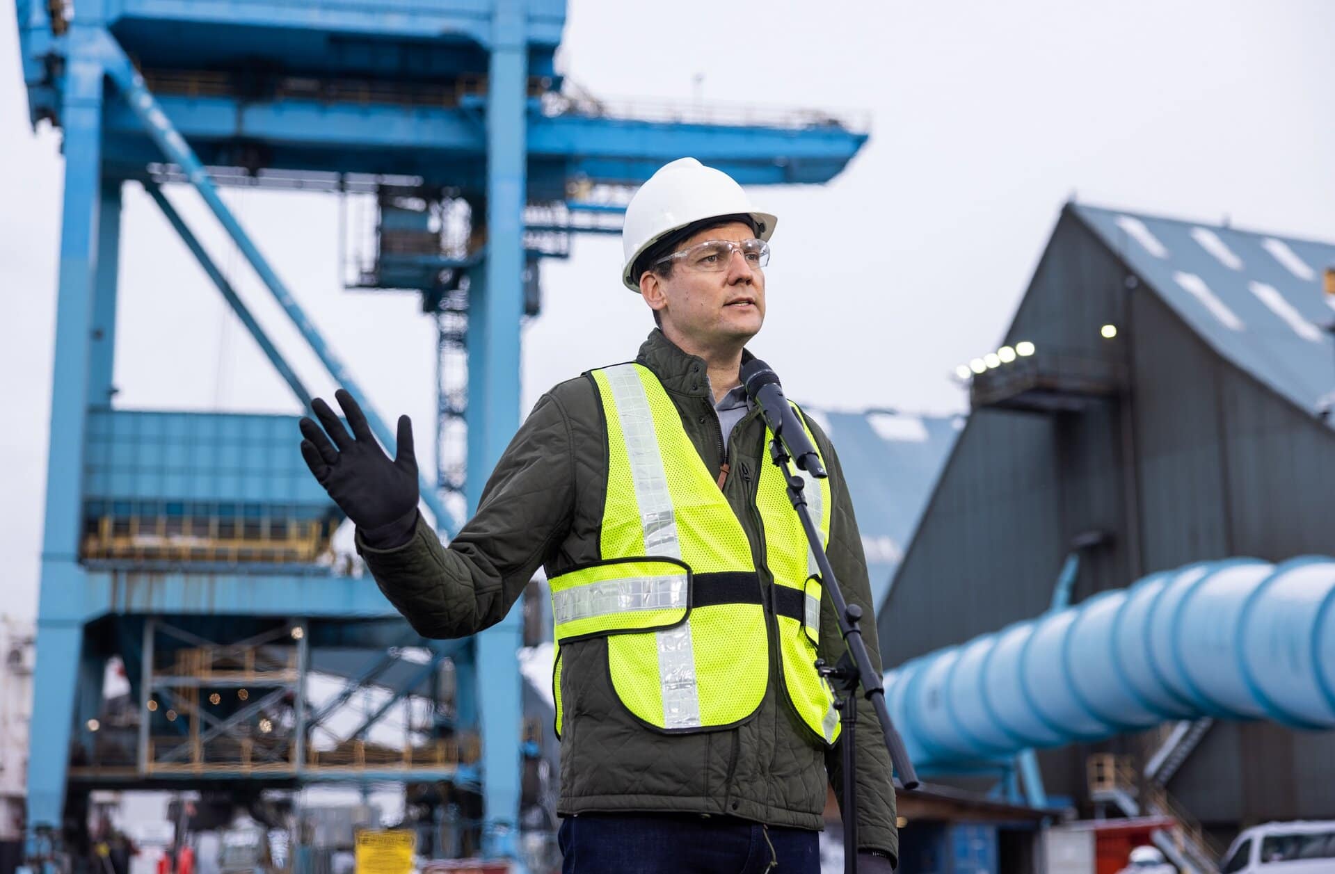 David Eby, wearing a hard hat, reflective vest and safety glasses, holds up a hand while speaking at a microphone with a towering industrial facility behind him
