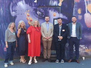 Six adults stand in a row outside Brighton School, smiling in front of a colorful mural featuring a painted face. Dressed in semi-formal attire, they represent the school’s commitment to challenging gender stereotypes.