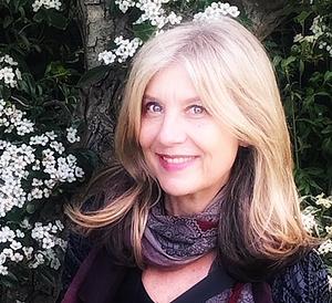 A woman with blonde hair smiles at the camera, standing outdoors in front of a blossoming tree with clusters of small white flowers. She wears a patterned scarf and a dark top, breaking free from traditional gender stereotypes.