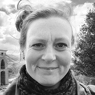A black and white close-up of a woman smiling outdoors. She has her hair up and is wearing a scarf. In the background, there's a tree and part of a building under a cloudy sky.
