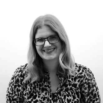 A person with long hair and glasses smiles at the camera. They are wearing a patterned blouse. The background is plain white. The image is in black and white.