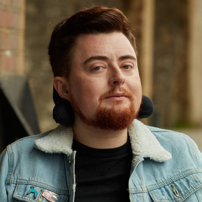 A picture of Jamie Hale against a blurred outdoor background. They are white, with dark red hair and a dark red beard. Their electric wheelchair headrest is behind their head, and they are wearing a blue denim jacket