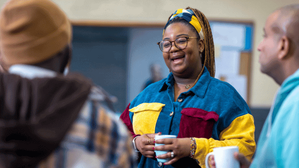 A person wearing glasses and a colorful jacket smiles while holding a mug, engaged in conversation with two others. They are indoors, suggesting a social setting.