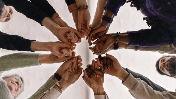 A group of diverse hands reaching towards the center, forming a circle against a light background. The hands are from different individuals, showcasing various skin tones and bracelets, symbolizing unity and collaboration.
