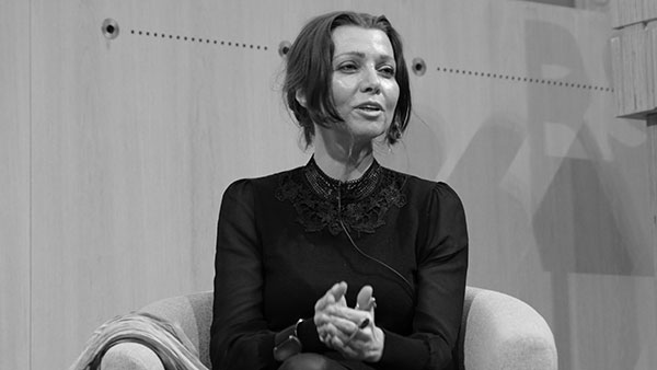 A woman with short hair is seated and speaking. She is wearing a long-sleeved, dark outfit with a detailed collar. The background consists of light-colored wooden panels with some text partially visible.
