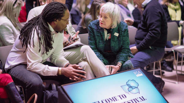 A man and woman sit and talk in a crowded venue. The man has long braids and glasses, while the woman has short white hair and a patterned jacket. In the foreground, a large screen reads "Welcome to the Festival.