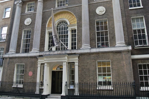 The exterior of the historic building, home to the Royal Society for the Arts, features tall columns and large windows adorned with decorative plaques. A flag waves proudly above the entrance, while a black wrought iron fence gracefully borders the sidewalk.