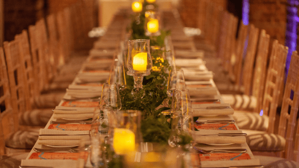 A long dining table elegantly set with folded napkins and glassware. Candlelit ambiance features glowing pillar candles in glass holders and a green leafy garland running along the center. Wooden chairs line both sides.