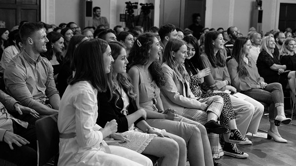 A black-and-white photo of an audience sitting in rows, focused on an event. People of various ages are smiling and clapping, suggesting they are enjoying the performance or presentation. The room is well-lit with a few visible cameras in the background.