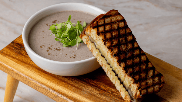 A bowl of creamy mushroom soup garnished with fresh greens sits on a wooden tray. A grilled cheese sandwich with grill marks is leaning against the bowl.