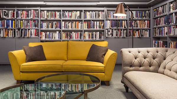 A cozy library setting with a yellow sofa and a beige tufted couch in front of bookshelves filled with colorful books. A modern floor lamp with a metallic shade stands beside the sofas, illuminating the glass coffee table.