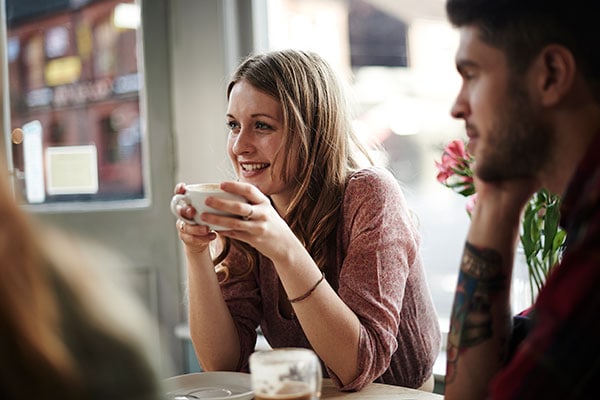 A woman smiles while holding a cup, sitting at a table in a cozy café. A blurred figure of a man is beside her, with tattoos visible on his arm. Sunlight filters through the window, creating a warm atmosphere.
