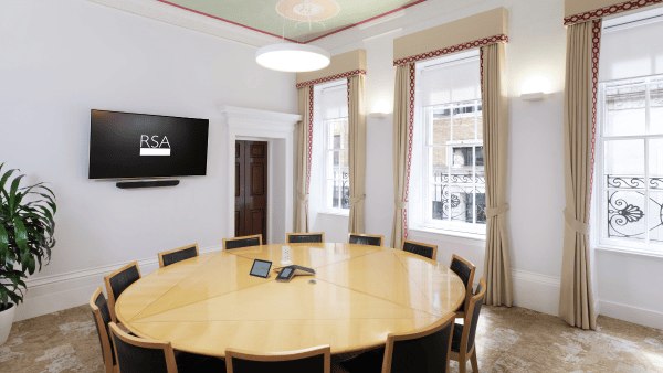 A meeting room with a large oval wooden table surrounded by ten chairs. A wall-mounted screen displaying "RSA" is on the left. There are two large windows with curtains, and a potted plant in the corner. A tablet lies on the table.