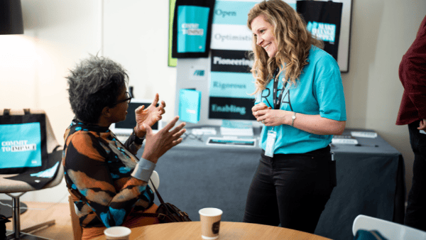 Two people talking at a table with coffee cups. One, wearing a colorful top, gestures with hands. The other, in a blue shirt with a lanyard, listens and smiles. A table with promotional materials is in the background.